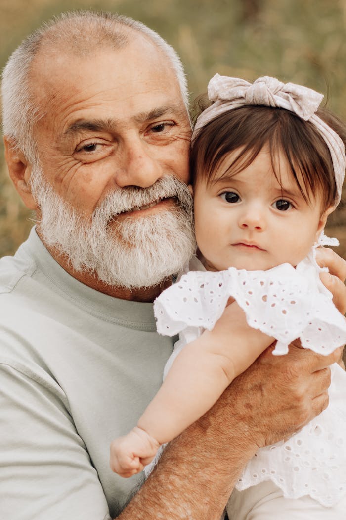 A loving grandfather embraces his baby granddaughter outdoors on a sunny day.